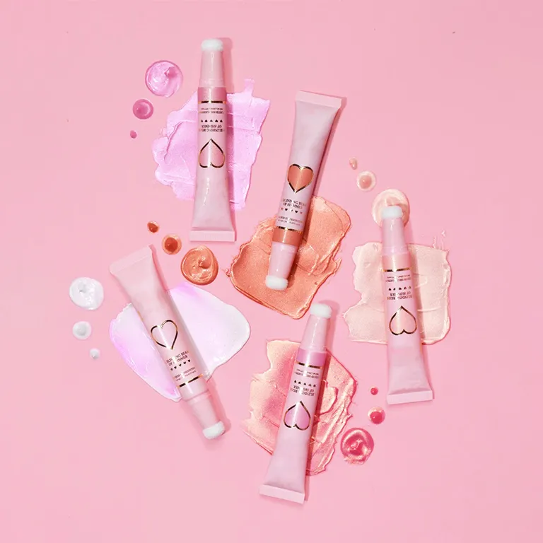 Five tubes of liquid highlighter with heart design on a pink background with makeup smears.