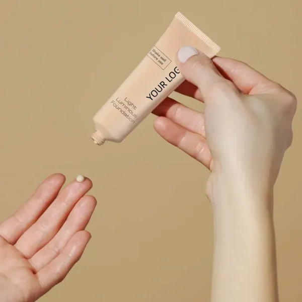 Close-up of a hand squeezing a drop of foundation from a tube labeled 'Your Logo' onto a finger.