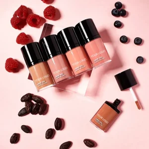 Five liquid blush bottles surrounded by raspberries, blueberries, and cocoa beans on a pink background.