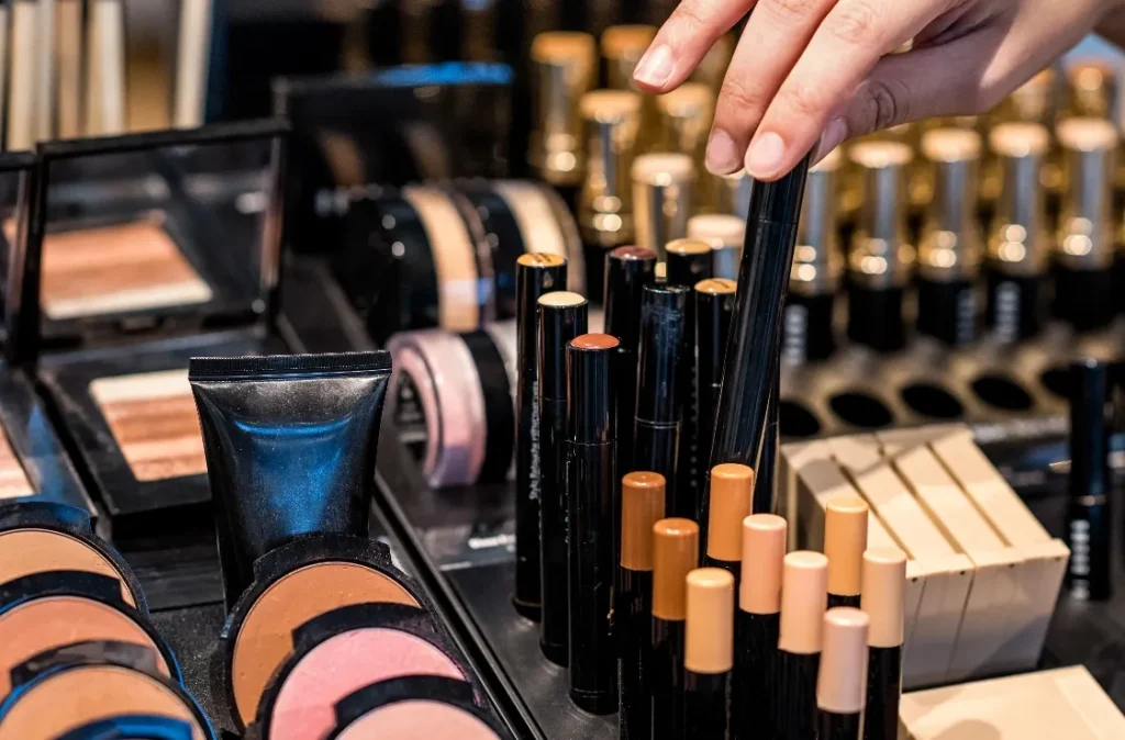 A hand reaching for a makeup product among various cosmetics displayed, including foundations, powders, and lip products.