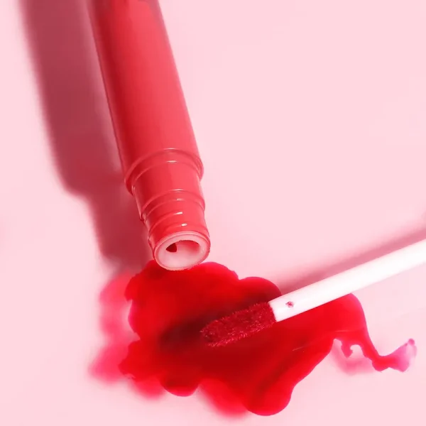 A red lip gloss spilled on a pink surface with an applicator brush.