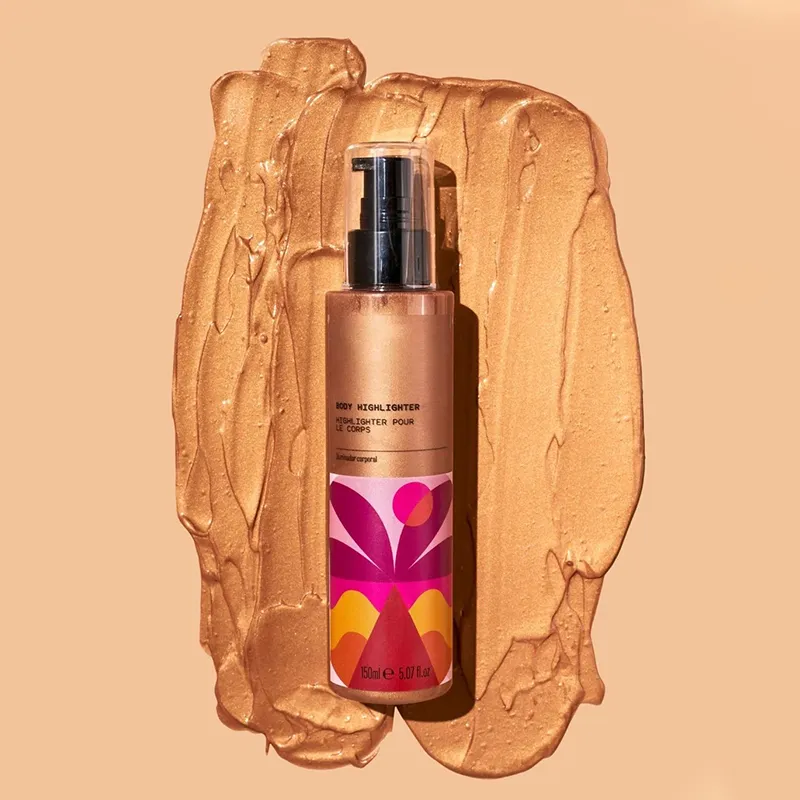 A bottle of body highlighter with a colorful label placed on a spread of golden cream.