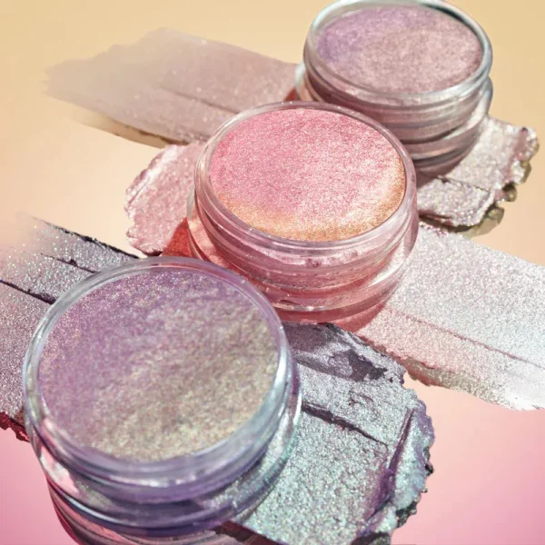 Image showcasing three small containers filled with shimmering cosmetic glitter in various shades including pink, purple, and silver, with additional glitter spread out behind them on a gradient background.