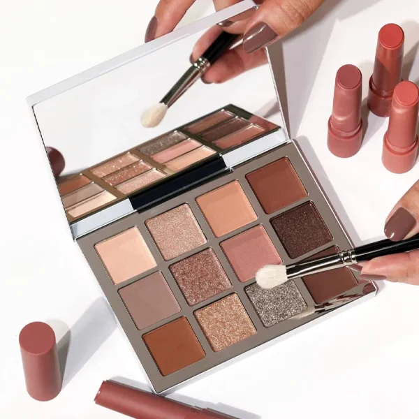 A flat-lay image of a makeup set featuring a 12-shade eyeshadow palette and multiple neutral-colored lipsticks. A hand holding an eyeshadow brush is also visible.