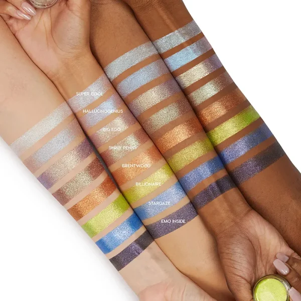 A display of eyeshadow swatches labeled with names like Super Cool, Hallucinogenus, Big Ego, Shiny Penny, Brentwood, Billionaire, Stargaze, and Emo Inside shown on arms of various skin tones.