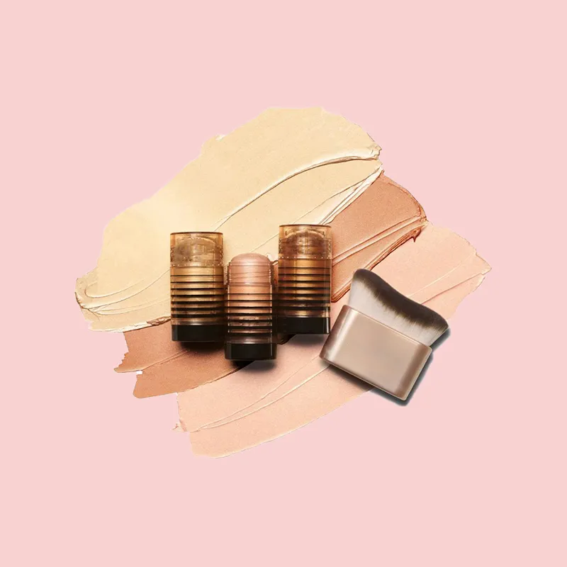 Three makeup sticks and a brush on smeared makeup swatches against a pink background.