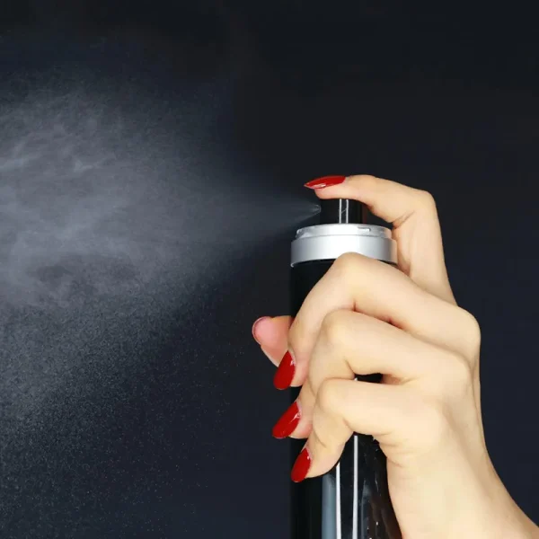 A hand with red nails is pressing the nozzle of a black spray can, releasing a fine mist against a dark background.