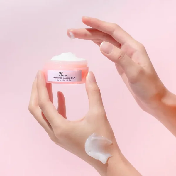 A jar of Xiran cherrywood cleansing balm being held by a hand, with product applied on the skin.