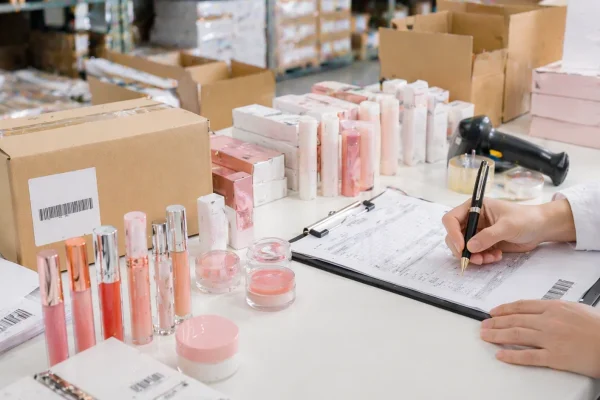 Cosmetic inventory management in warehouse Person managing cosmetics inventory with boxes and products on a table, writing on a clipboard in a warehouse setting.