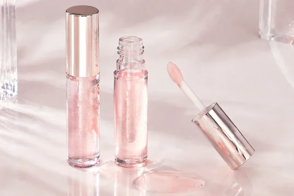 Pink lip gloss with shiny packaging Open and closed bottles of pink lip gloss with a shiny applicator and reflective packaging on a light surface.