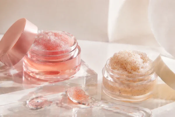 Two jars of lip scrubs in natural light Two small jars of lip scrubs, one pink and one beige, placed on a clear surface with glass elements around them.